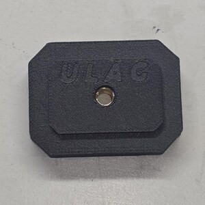 May include: A black rectangular object with rounded corners, featuring the embossed text "ULAC" and a central hole with a brass-colored insert. The object is set against a light gray surface.