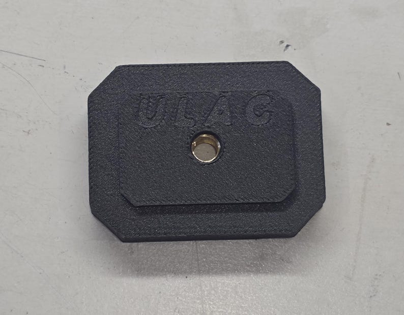 May include: A black rectangular object with rounded corners, featuring the embossed text "ULAC" and a central hole with a brass-colored insert. The object is set against a light gray surface.