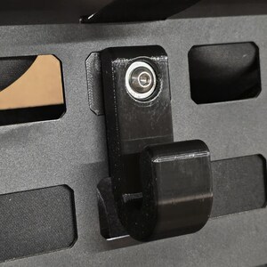 May include: A black, 3D-printed hook attached to a dark gray panel with rectangular cutouts. The hook has a curved design and is secured with a silver fastener. The panel has a textured surface and is likely part of a storage or mounting system.