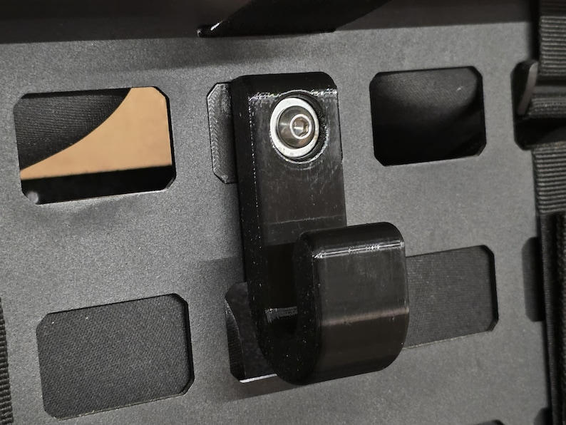 May include: A black, 3D-printed hook attached to a dark gray panel with rectangular cutouts. The hook has a curved design and is secured with a silver fastener. The panel has a textured surface and is likely part of a storage or mounting system.