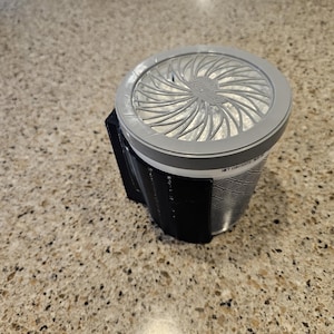 May include: A gray and black air freshener with a circular design on the top. The air freshener is attached to a wall mount.