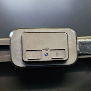 May include: A gray rectangular button with two smaller buttons and a blue light. The buttons are labeled with arrows.