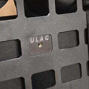 May include: Close-up of a black metal panel with a grid of rectangular cutouts. A small, rectangular plate with the letters "ULAG" is affixed to the panel. The plate has a threaded hole in the center.