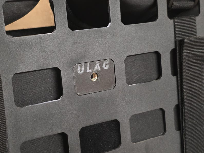 May include: Close-up of a black metal panel with a grid of rectangular cutouts. A small, rectangular plate with the letters "ULAG" is affixed to the panel. The plate has a threaded hole in the center.