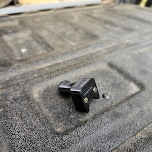 May include: A black plastic truck bed tie-down anchor with two metal hooks. The anchor is designed to be used with a rope or strap to secure cargo in the bed of a truck.