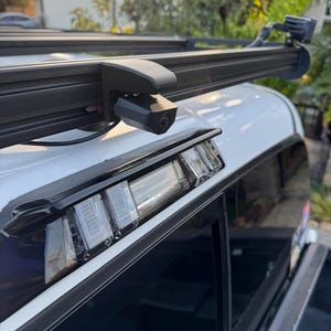 May include: A black roof rack system with a mounted camera and LED light bar on a white vehicle. The camera is positioned on the side of the rack, and the LED light bar is at the rear. The vehicle's rear window and brake lights are also visible.