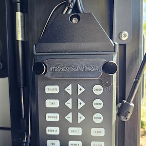 May include: A black control panel with buttons for various functions, including tailgate, tent, awning, back light, side light, light bar, fan, heater, and 12V outlet. The panel has a black and silver logo that says "Mountain Made" and two black knobs.