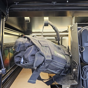 May include: A black and green tool bag hanging from a hook inside a vehicle. The bag has a zipper and a strap. The bag is hanging from a hook attached to a metal bar.