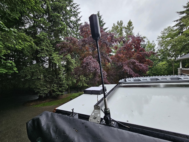 K&ouml;nnte beinhalten: Eine schwarze Antenne, die auf einer wei&szlig;en Oberfl&auml;che, wahrscheinlich einem Fahrzeugdach, montiert ist, mit einem Hintergrund aus &uuml;ppigen gr&uuml;nen und roten B&auml;umen unter einem bew&ouml;lkten Himmel. Die Antenne ist hoch und schlank, mit einem rechteckigen Oberteil.