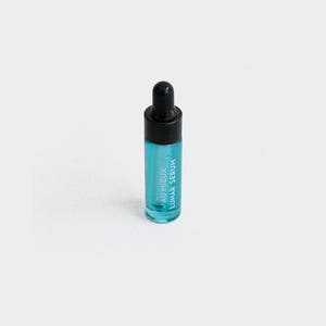 Op de afbeelding: Een klein, doorzichtig glazen flesje met een zwarte druppelaar. Het flesje is gevuld met een lichtblauwe vloeistof en heeft de tekst "AU MIEUX LUNAR SERUM" op de zijkant.