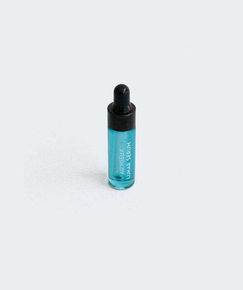 Op de afbeelding: Een klein, doorzichtig glazen flesje met een zwarte druppelaar. Het flesje is gevuld met een lichtblauwe vloeistof en heeft de tekst "AU MIEUX LUNAR SERUM" op de zijkant.