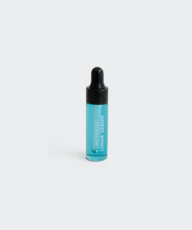 Op de afbeelding: Een klein, doorzichtig glazen flesje met een zwarte druppelaar. Het flesje bevat een lichtblauwe vloeistof en heeft de tekst "AU MIEUX LUNAR SERUM" op de zijkant.
