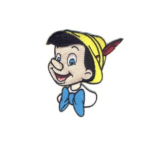 Pode incluir: Etiqueta bordada com uma imagem de desenho animado do Pinóquio, um fantoche de madeira com um nariz grande, usando um chapéu amarelo com uma pena vermelha e uma gravata borboleta azul.