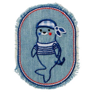 Könnte beinhalten: Gestickter Denim-Patch mit einem Cartoon-Robben in Matrosen-Outfit. Die Robbe trägt ein blau-weiß gestreiftes Hemd und eine Matrosenmütze. Der Patch hat einen ausgefransten Rand und einen rot-blauen Rand.