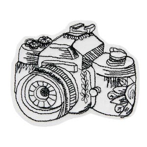 Può includere: Toppa ricamata con un design di fotocamera in bianco e nero. La toppa mostra cuciture dettagliate, tra cui l'obiettivo, il flash e i pulsanti. La fotocamera è su uno sfondo bianco, evidenziando l'artigianato.