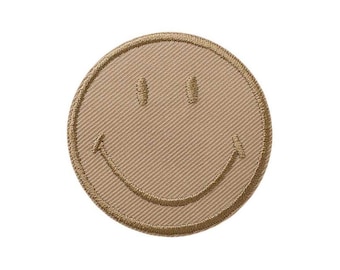 Op de afbeelding: Een ronde geborduurde patch in een effen beige kleur met een gestikt smiley-gezicht. De patch heeft een dikke, verhoogde rand en twee kleine verticale ovale ogen met een brede, gebogen mondlijn, allemaal uitgevoerd in bijpassend beige garen.