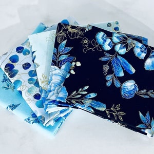 Könnte beinhalten: Fünf gefaltete Stoffquadrate mit floralen Mustern in Blau- und Goldtönen. Die Stoffe weisen verschiedene Designs auf, darunter Blumen und Blätter, vor hellblauen und marinefarbenen Hintergründen. Die Anordnung zeigt die verschiedenen Muster und Farben.
