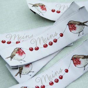 May include: White ribbon with a repeating pattern of red berries, a robin, and the text "Meins" in a light brown font.