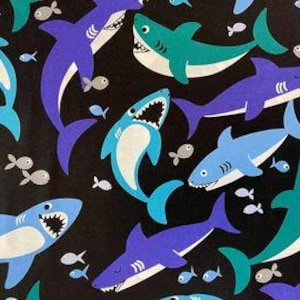 Op de afbeelding: Een zwarte stof met een herhalend patroon van blauwe, turquoise en paarse haaien met witte tanden en witte buiken. Kleine grijze vissen zijn verspreid over het patroon.