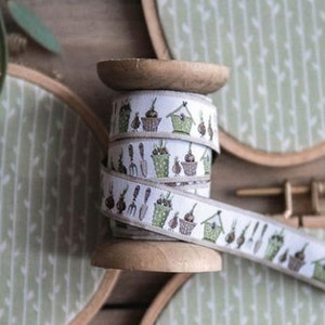 Peut inclure: Une bobine de ruban blanc avec un motif répété vert et marron d'outils de jardinage, de pots de fleurs et de bulbes. Le ruban est enroulé autour d'une bobine en bois.