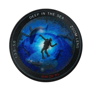 Può includere: Toppa rotonda con un subacqueo in un mare blu profondo, circondato da squali. Il design imita un obiettivo fotografico, con le scritte "DEEP IN THE SEA" e "ZOOM LENS" sul bordo. L'obiettivo ha le misure 1:3.5 - 5.6 e 14-42 mm.