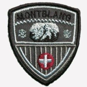 Può includere: Una toppa ricamata grigia con le parole "MONTBLANC" in lettere bianche. La toppa presenta una catena montuosa stilizzata con una croce bianca su sfondo rosso.