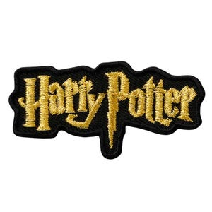 Könnte beinhalten: Ein gestickter Aufnäher mit dem Schriftzug Harry Potter in einer stilisierten goldenen Schriftart. Die Buchstaben sind mit metallischem Goldfaden auf einem schwarzen Stoffhintergrund gestickt, mit einem schwarzen Rand.