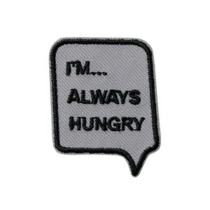 Op de afbeelding: Een grijs en zwart geborduurd strijk-op-patch met een spraakballonontwerp. De tekst in de spraakballon luidt "I'm... ALWAYS HUNGRY".
