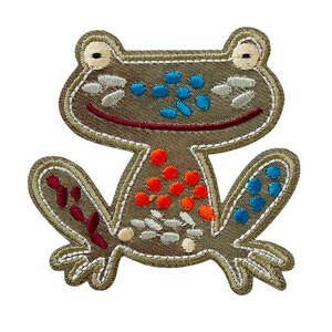 Peut inclure: Un patch brodé de grenouille vert avec des détails rouges, orange, bleus et blancs. La grenouille sourit et a une surface en tissu texturée.