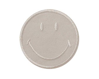 Op de afbeelding: Een ronde geborduurde patch in een neutrale beige kleur met een gestikt smiley-gezicht. De patch heeft een verhoogde, textuurrijke rand en twee kleine verticale ovale ogen boven een brede, gebogen mond, allemaal uitgevoerd in bijpassend garen.