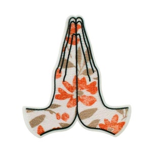 Peut inclure: Écusson brodé représentant des mains jointes en prière. Le patch a un fond blanc avec un motif floral orange et marron. Le contour des mains est noir.