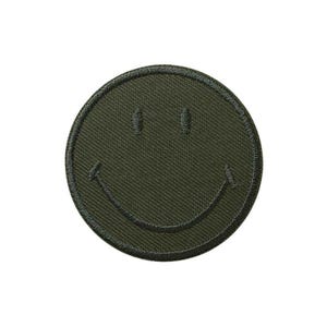 Könnte beinhalten: Runder, olivgrüner, gestickter Aufnäher mit einem lächelnden Gesicht. Der Aufnäher hat einen gestickten Rand und ein minimalistisches Design. Das Smiley-Gesicht hat zwei kleine, gestickte Augen und einen gebogenen Mund.