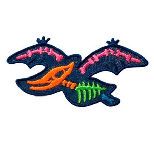 Peut inclure: Un patch brodé bleu foncé représentant une chauve-souris volante avec des détails roses, orange et verts. La chauve-souris a un design squelettique avec un crâne et des os.