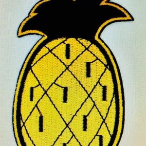 Op de afbeelding: Een gele ananas met een zwarte omtrek en een zwarte bovenkant. De ananas is geborduurd met een ruitpatroon.