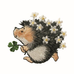 Könnte beinhalten: Gestickter Aufnäher eines braunen und schwarzen Igels mit weißen Blüten auf dem Rücken, der ein grünes vierblättriges Kleeblatt hält.