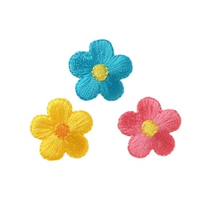 Könnte beinhalten: Drei gestickte Blumenpatches in Blau, Gelb und Rosa. Jede Blume hat eine gelbe Mitte.