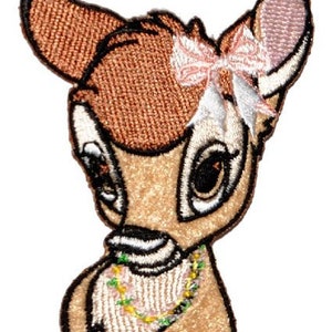 Könnte beinhalten: Gestickter Aufnäher mit einem Cartoon-Hirsch mit rosa Schleife und Perlenkette. Der Hirsch hat braunes Fell, ein weißes Gesicht und schwarze Umrisse. Der Aufnäher ist für das Aufnähen auf Kleidung oder Accessoires gedacht.