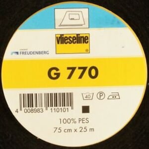 Può includere: Un rotolo di interfodera termoadesiva nera Vlieseline G 770. L'etichetta è gialla con testo nero e un codice a barre bianco. L'etichetta include anche un'icona di istruzioni di lavaggio, un simbolo per un'impostazione di stiratura permanente e due icone di ferro da stiro. L'etichetta indica "100% PES" e "75 cm x 25 m".
