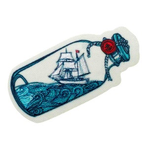 Puede incluir: Un parche de tela blanco con una imagen bordada de un barco en una botella. La botella es azul y blanca con una rosa roja en la parte superior. El barco es blanco con velas azules y una bandera roja.
