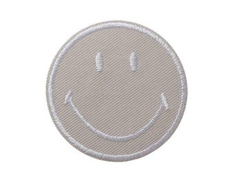 Op de afbeelding: Een cirkelvormige geborduurde patch met een lichtbeige achtergrond en een wit gestikt smiley-gezicht. De patch heeft een strakke, ronde rand en een zichtbare diagonale stoftextuur over het gehele oppervlak van het ronde embleem.