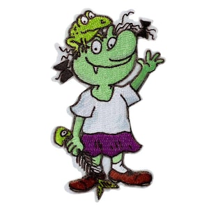 Può includere: Una toppa ricamata da stirare con un personaggio dei cartoni animati verde con una rana sulla testa, che indossa una camicia bianca e una gonna viola, tenendo un bastone verde con una piuma all'estremità.