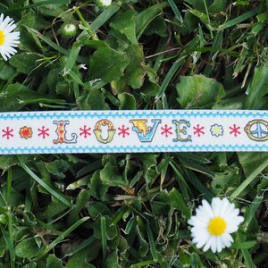 May include: A white ribbon with a blue border and the word "LOVE" in colorful letters. The ribbon is decorated with peace signs, four-leaf clovers, and flowers. The ribbon is lying on a bed of green grass.