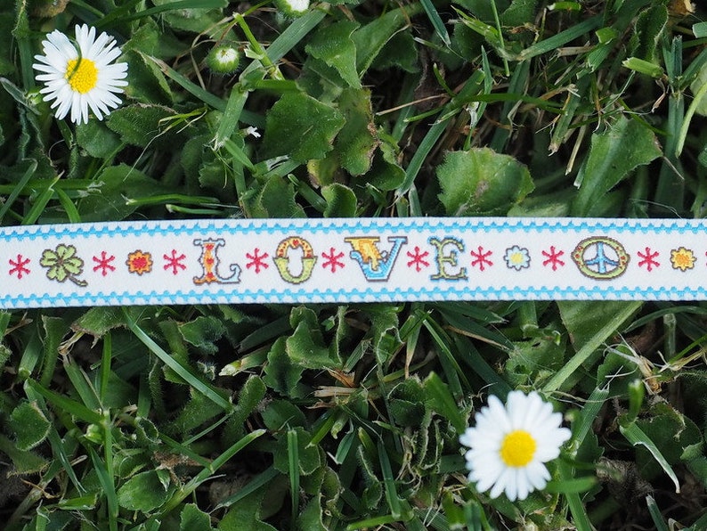 May include: A white ribbon with a blue border and the word "LOVE" in colorful letters. The ribbon is decorated with peace signs, four-leaf clovers, and flowers. The ribbon is lying on a bed of green grass.