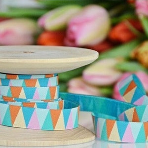 Peut inclure: Un bobine de ruban à motifs turquoise, orange, rose et crème avec un motif géométrique de triangles. Le ruban est enroulé autour d'une bobine en bois.