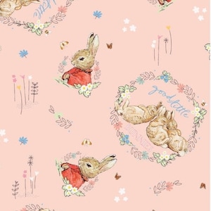 Peut inclure: Un tissu rose avec un motif répété de lapins. Les lapins sont dans différentes poses, y compris assis, dormant et en train de se câliner. Le tissu présente également des couronnes de fleurs avec le texte "good little bunnies" et "little bunnies".