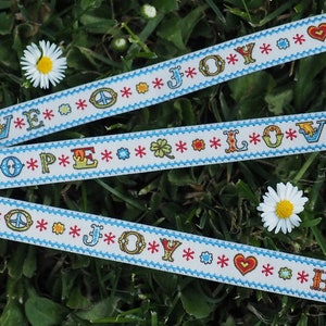 May include: Three white ribbons with blue trim and colorful embroidered words: "LOVE", "HOPE", and "JOY". The ribbons are decorated with peace signs, hearts, and stars.