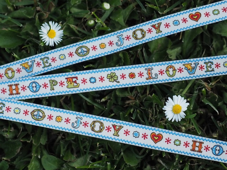 May include: Three white ribbons with blue trim and colorful embroidered words: "LOVE", "HOPE", and "JOY". The ribbons are decorated with peace signs, hearts, and stars.