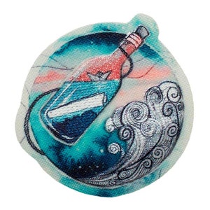 Könnte beinhalten: Ein runder, gestickter Patch mit einer blauen Glasflasche mit einem roten Korken und einem weißen Papier darin. Die Flasche schwebt auf einer türkisfarbenen Welle mit einem weißen Wirbelmuster. Der Hintergrund ist ein hellblauer und rosafarbener Aquarell-Wasch.