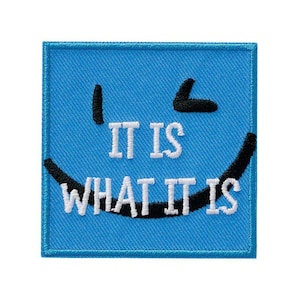 Op de afbeelding: Een blauwe geborduurde patch met een lachend gezicht en de tekst "It is what it is".