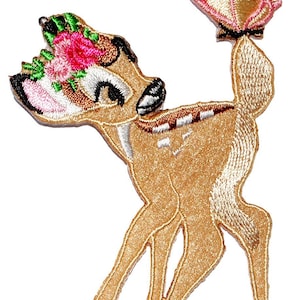 Op de afbeelding: Geborduurde strijkpatch met een cartoon-hert met een roze bloemenkrans en een vlinder op zijn staart. Het hert is bruin met witte accenten en heeft grote, expressieve ogen.